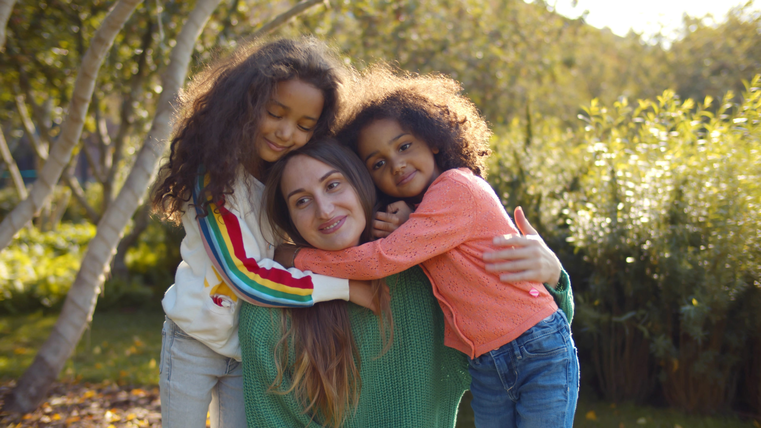 Smiling white woman and her two biracial children hugging her in a park.