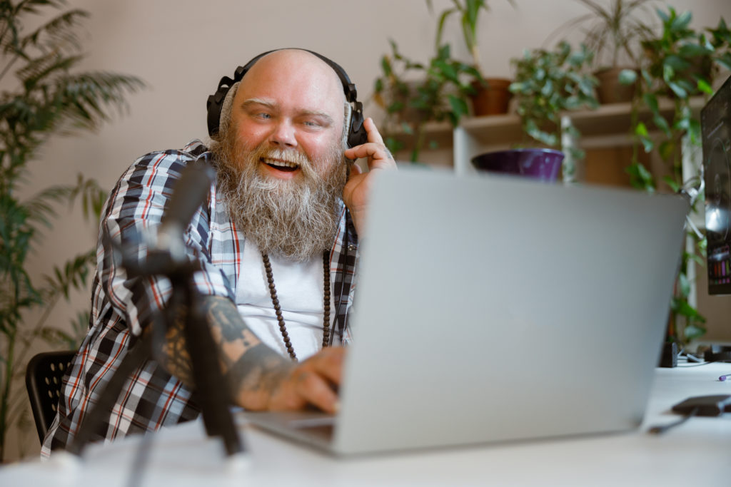 Man with a long gray beard, laughing while using a laptop, and wearing gaming headphones and using a mic.