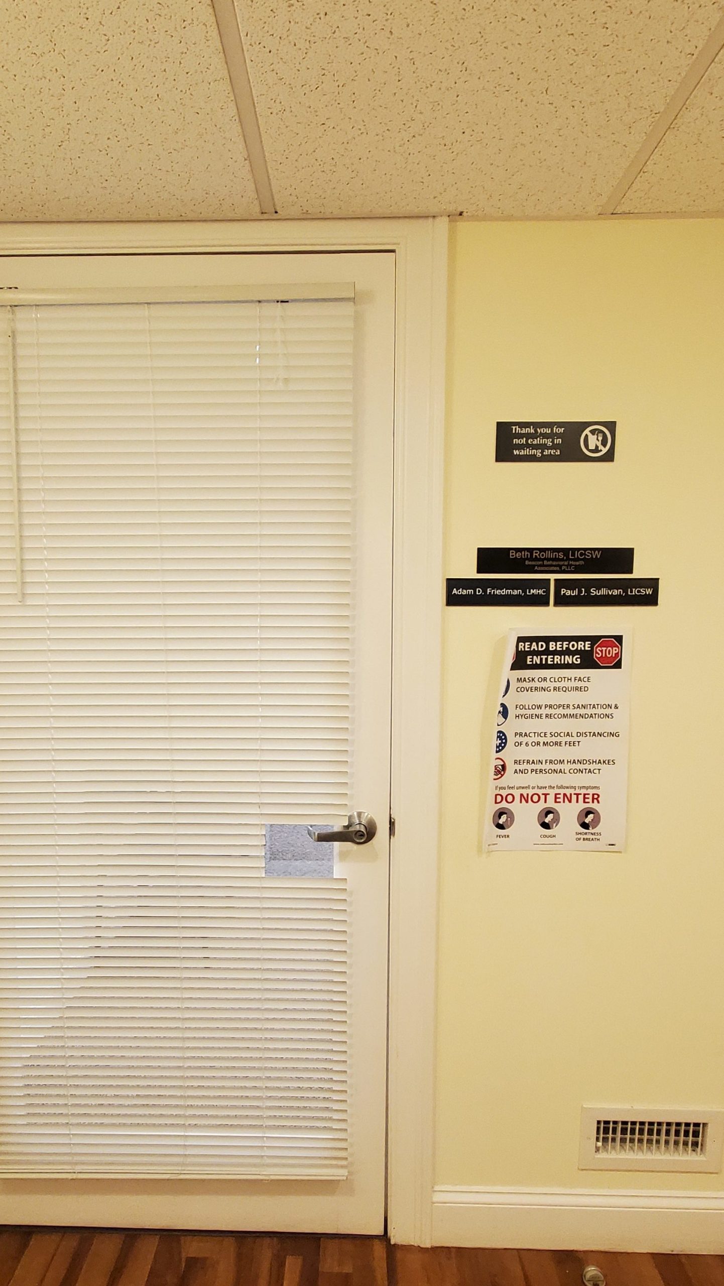 The door to the waiting room. The door has a glass window in it and white blinds prevent people from looking inside the room. On the right hand side are three name plates, a sign asking people to not eat in the waiting area, and a sign asking people to wear face masks. 