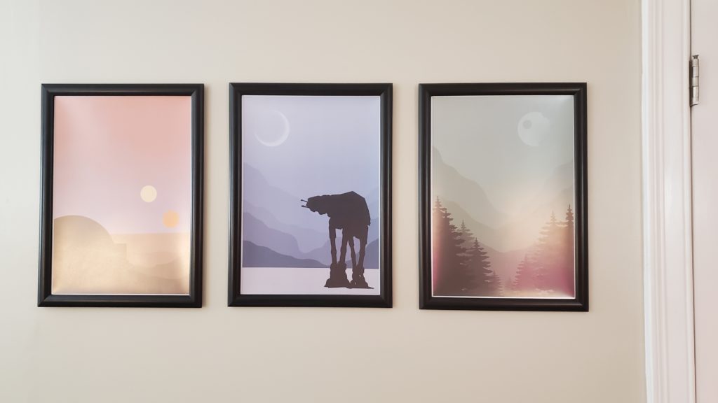 Three poster prints of scenes from Star Wars. From left to right: a minimalist pinkish red illustration of Tatooine with a moisture farm, a minimalist blue illustration of Hoth with an AT-AT in the foreground, and a minimalist green illustration of Endor with the Death Star 2 in the upper right corner.