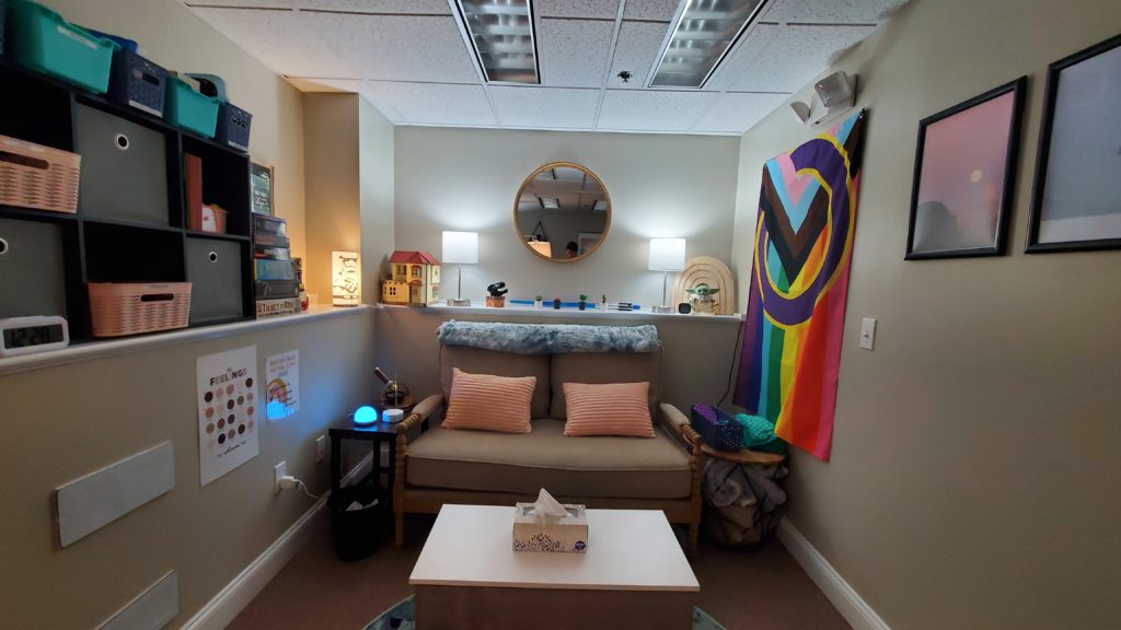 Therapy office with dim lighting. A love seat is against the far wall with soft pillos and a soft blanket on it. An inclusive pride flag is hanging on the right side wall. The lighting is dim and calming. 
